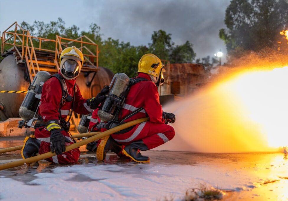 Firefighters in red suits and helmets extinguish a fire with a hose, surrounded by smoke and industrial equipment.