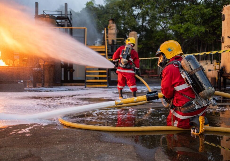 Firefighters in red suits and yellow helmets spray water from hoses to extinguish a fire in an industrial area, with visible flames and equipment in the background.